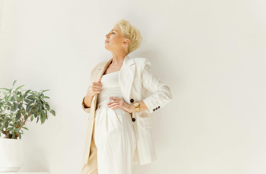short-haired woman wearing a long white coat and pants, with one hand wrapped around her waist