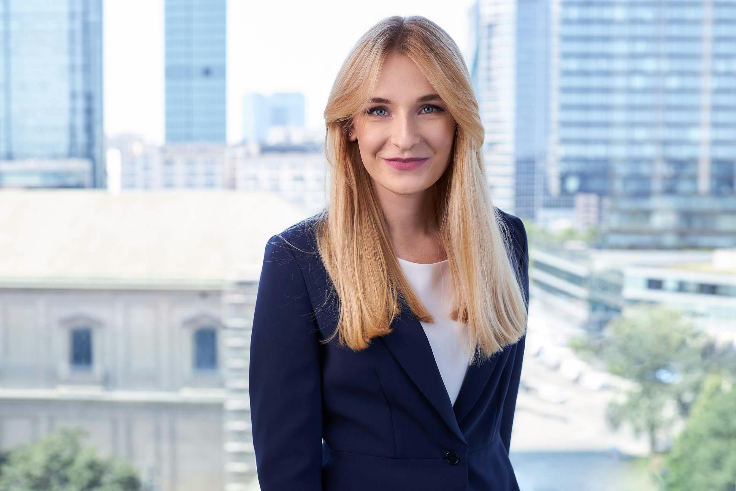 professional headshot of a blonde woman wearing a black blazer over a white top, with a city skyline in the background